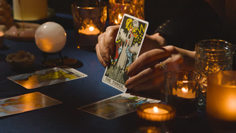Nahaufnahme-Einer-Frau,-Die-Auf-Einem-Tisch-Bei-Kerzenschein-Tarotkarten-Liest-Und-Die-Turmkarte-Hält
